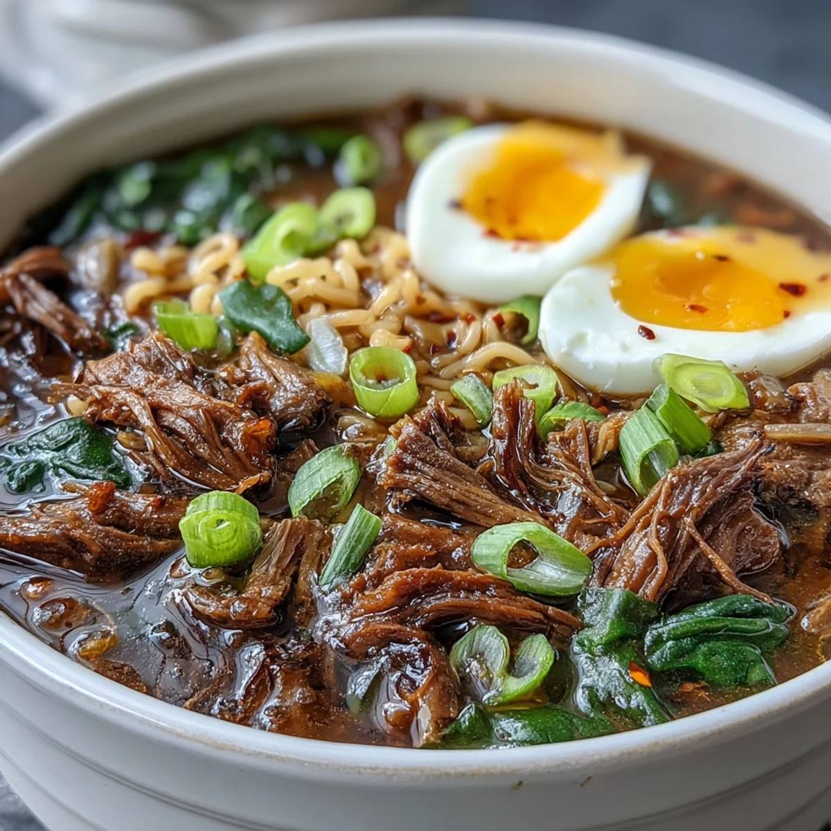 Savory slow cooker beef ramen noodles topped with chili oil, sesame seeds, and fresh spinach in steaming broth.